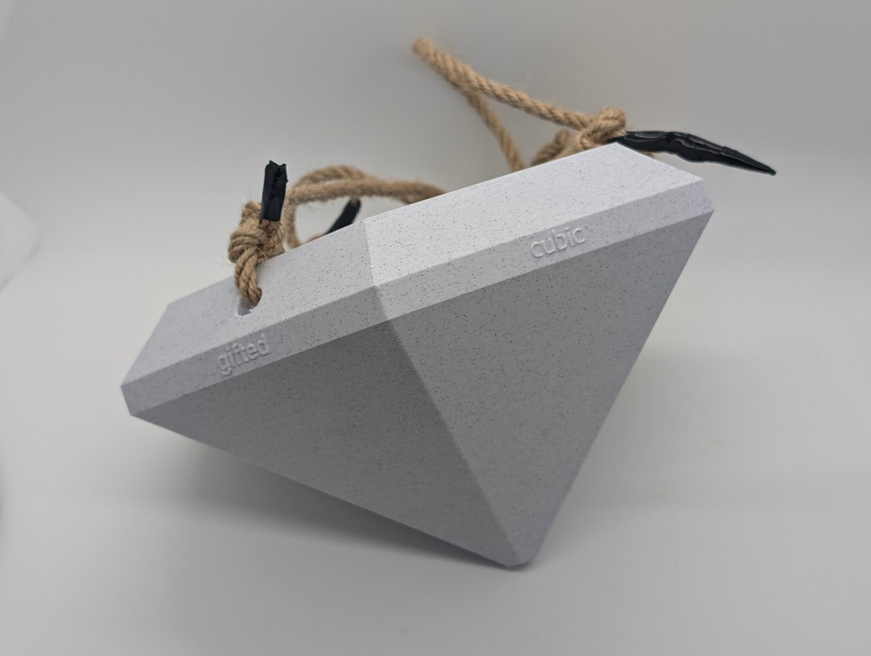 A close-up photograph of a large, 3D-printed diamond-shaped object positioned on its side against a plain white background. The object is light gray with a speckled texture and features embossed text on its facets that reads 'gifted cubic.' The diamond is secured with a rope harness, with black clips attached at two points. The image focuses on the geometric design, texture, and detailed craftsmanship of the 3D-printed object.