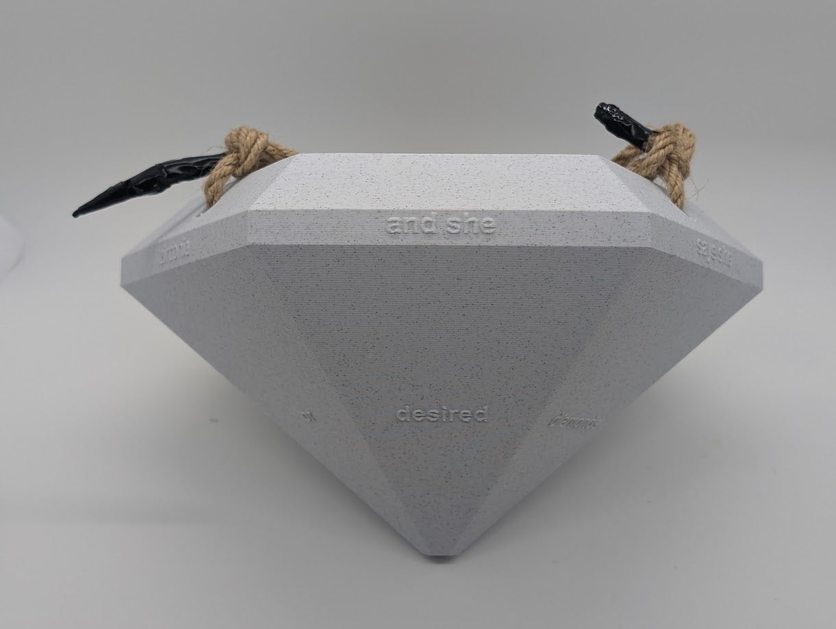 A close-up photograph of a large, 3D-printed diamond-shaped object placed on its side against a plain white background. The object is light gray with a speckled texture and features embossed text on its facets that reads 'and she desired diamonds.' The diamond is secured with a rope harness, with black clips attached at two points. The image highlights the geometric design, texture, and craftsmanship of the 3D-printed object.