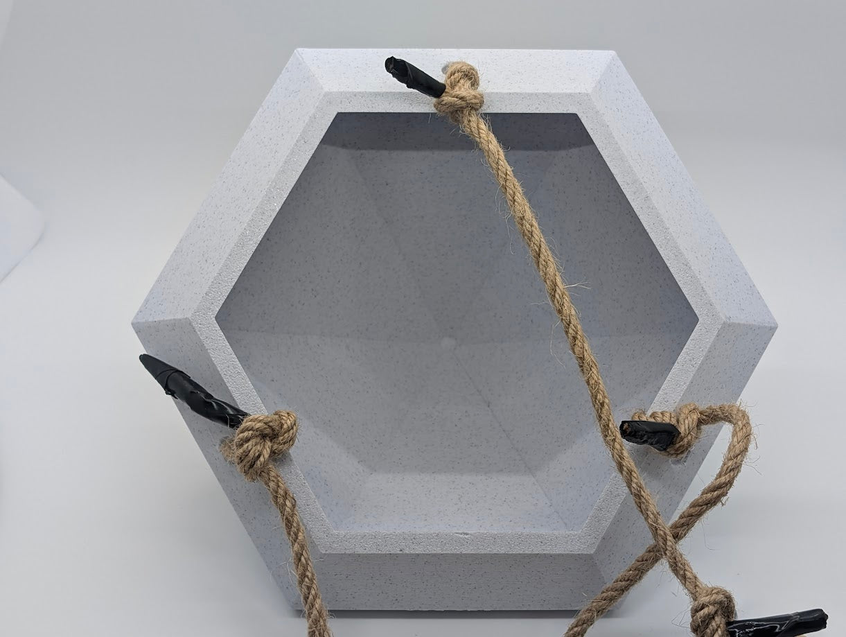 A photograph of a large, 3D-printed geometric object with an open, hexagonal interior, resembling a faceted gemstone or container. The object is light gray with a textured surface and features a rope harness for hanging. The rope is secured with black clips at multiple points on the object, providing stability for hanging. The object is positioned upright against a plain white background, emphasizing the precise angles and design of the 3D print.