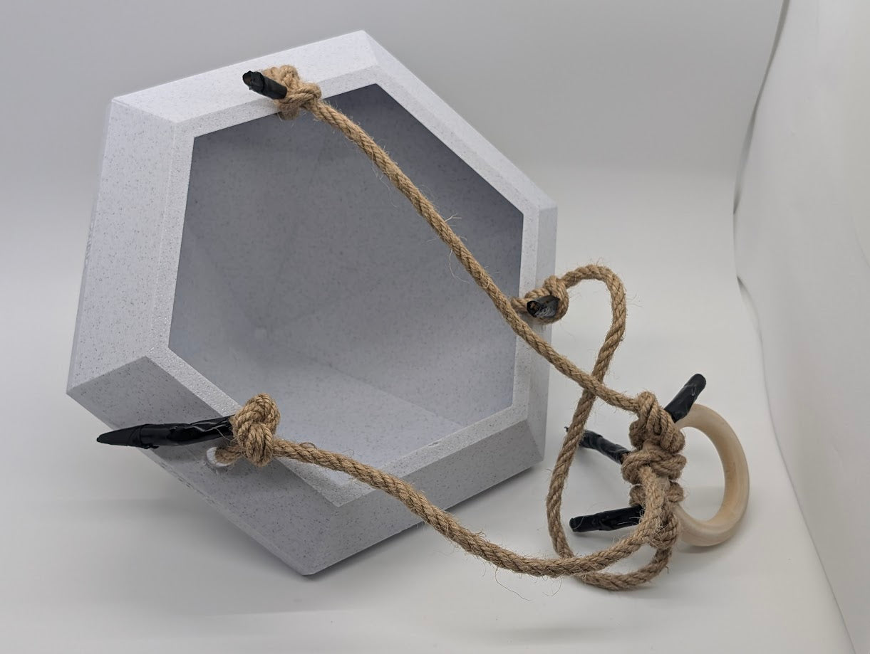 A photograph of a large, 3D-printed geometric object with an open, hollow interior, resembling a faceted gemstone or diamond. The object is light gray with a textured surface and is equipped with a rope harness for hanging. The rope is tied securely around the object and attached to a wooden ring, with black clips used for additional support. The object is placed on its side against a plain white background, highlighting the craftsmanship and functional design of the 3D print.