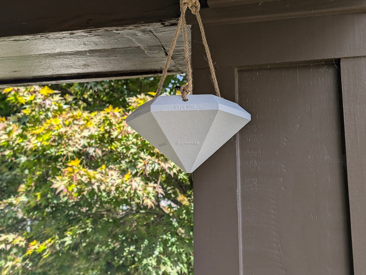 A photograph of a large, diamond-shaped 3D-printed object hanging from a rope under the eaves of an outdoor structure. The gray diamond features embossed text on its facets that reads 'says she never gifted diamonds and desired cubic zirconia.' The object is suspended against a background of green foliage, partially framed by the wooden structure. The scene highlights the contrast between the industrial design of the 3D-printed diamond and the natural outdoor environment.