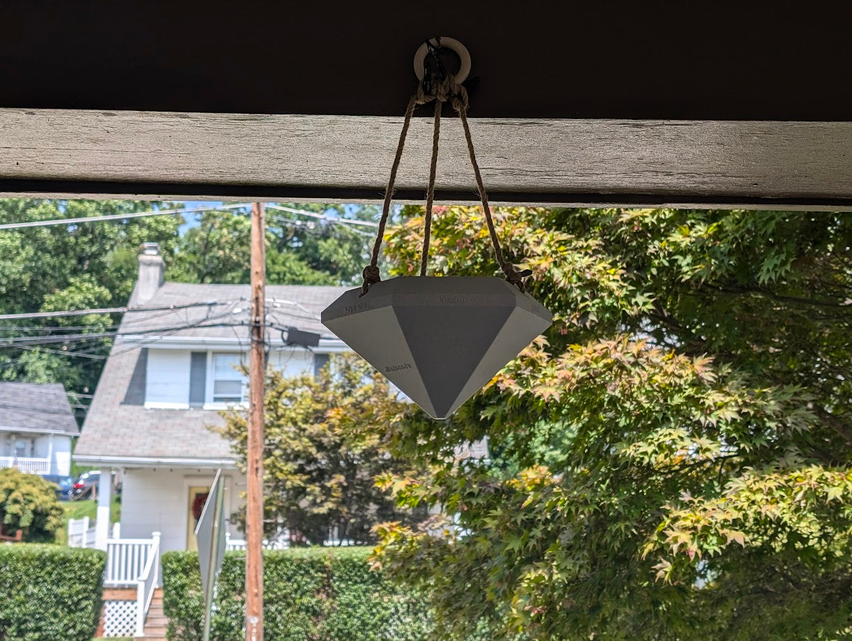 A photograph of a large, diamond-shaped 3D-printed object hanging from a rope under the roof of an outdoor structure. The gray diamond is suspended in front of a suburban neighborhood scene, with a house, trees, and power lines visible in the background. The diamond features embossed text on its facets, but the text is less visible due to the distance. The contrast between the industrial design of the 3D-printed diamond and the residential environment creates an intriguing visual juxtaposition.