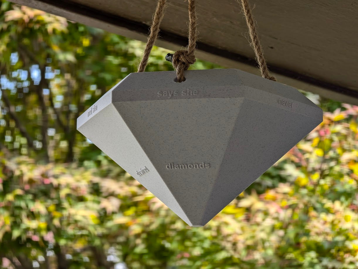 A photograph of a large, diamond-shaped 3D-printed object hanging from a rope under an outdoor structure. The object is gray with a matte finish and has embossed text on its facets that reads 'says she never gifted diamonds and desired cubic zirconia.' The diamond is suspended in front of a blurred background of greenery, creating a contrast between the geometric shape and the natural environment. The scene combines industrial design with a natural setting, highlighting the craftsmanship and creativity of t