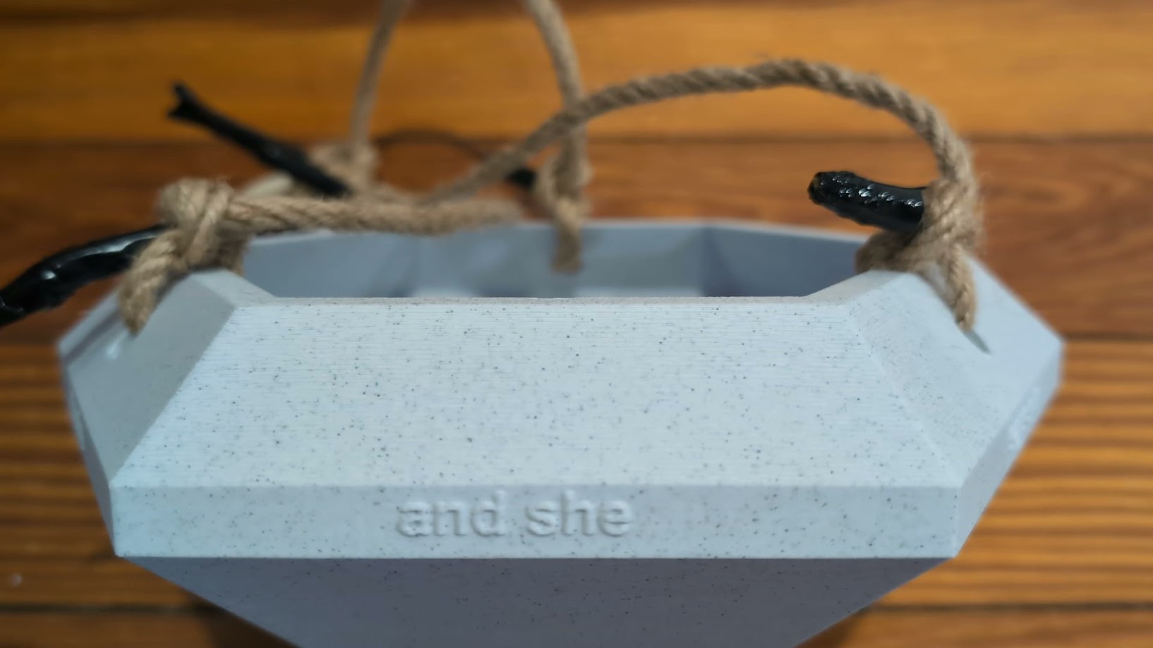 A close-up photograph of the top edge of a large, 3D-printed diamond-shaped object with a speckled gray texture. The focus is on the facet that features the embossed text 'and she.' The object is secured with a rope harness, with black clips visible where the rope is tied. The background shows a wooden surface, adding warmth and contrast to the industrial look of the 3D-printed object.