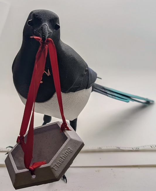 A close-up photograph of a black-and-white magpie figurine holding a red ribbon in its beak. The ribbon is attached to a small, 3D-printed, hexagonal object with the word 'Flashing' embossed on one of its sides. The figurine is positioned against a plain white background, emphasizing the contrast between the vivid colors of the bird's tail and the muted tones of the 3D-printed object. The scene captures a playful and artistic display, combining elements of nature and 3D printing.