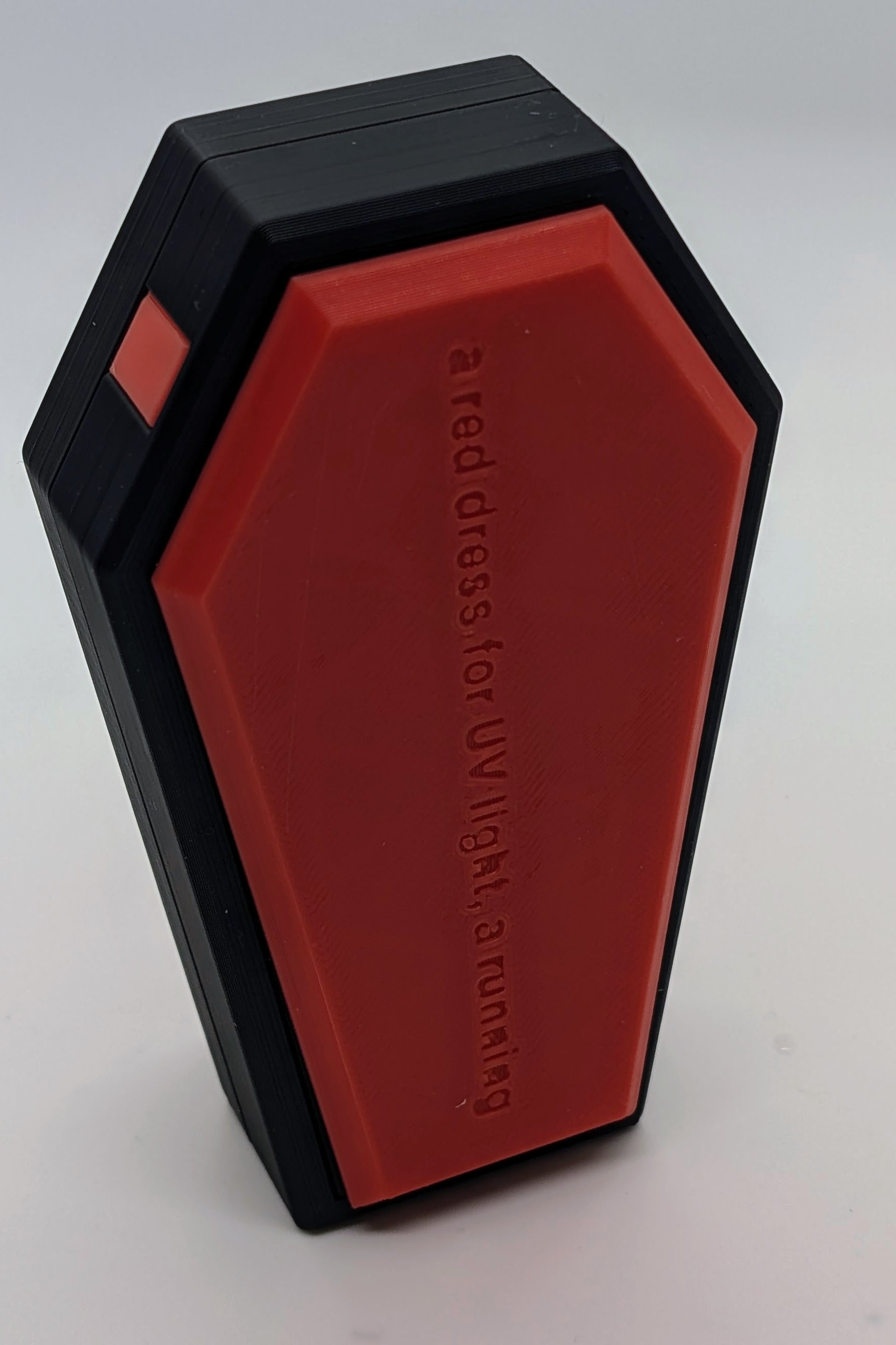 A photograph of a red and black 3D-printed object shaped like a coffin, standing upright against a plain white background. The red face of the object features embossed text that reads, 'a red dress, for UV light, a running.' The object has a sleek, angular design with sharp edges, showcasing the precision of the 3D printing process. The minimalist and modern aesthetic of the object makes it suitable for design-focused or creative projects.