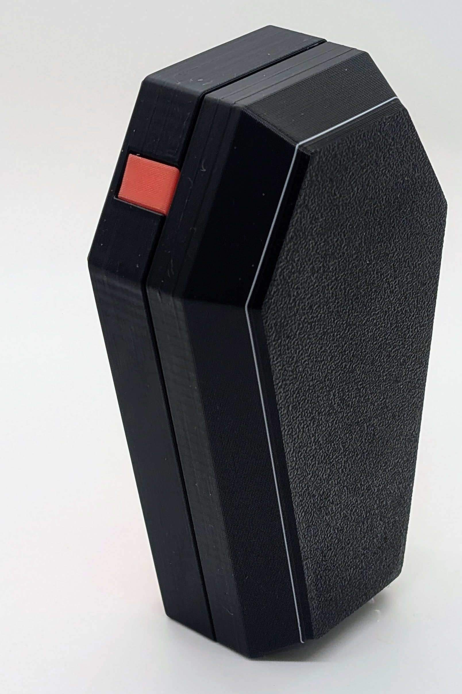 A photograph of a black, coffin-shaped 3D-printed object standing upright against a plain white background. The object features a textured surface and a small red button or detail on one side, adding a subtle contrast to the sleek, angular design. The minimalist aesthetic and sharp edges highlight the precision and quality of the 3D printing process.