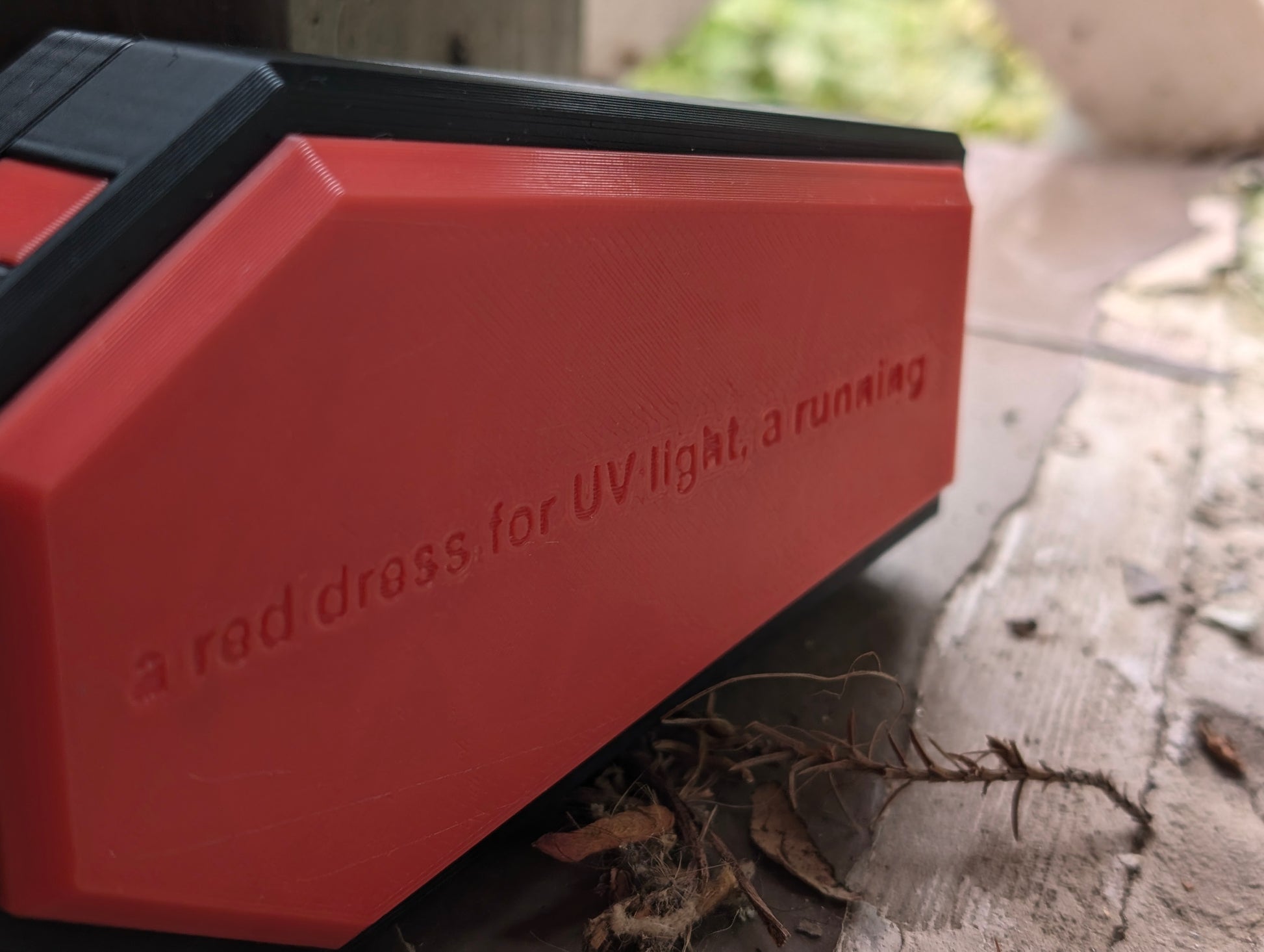 A close-up photograph of a red, 3D-printed object with embossed text that reads, 'a red dress, for UV light, a running.' The object has sharp, angular edges and appears to be partially resting on a tiled floor, with some dried leaves nearby. The background is slightly blurred, focusing attention on the texture and detail of the 3D print. The scene combines industrial design with a natural, weathered environment, highlighting the precision and craftsmanship of 3D printing.