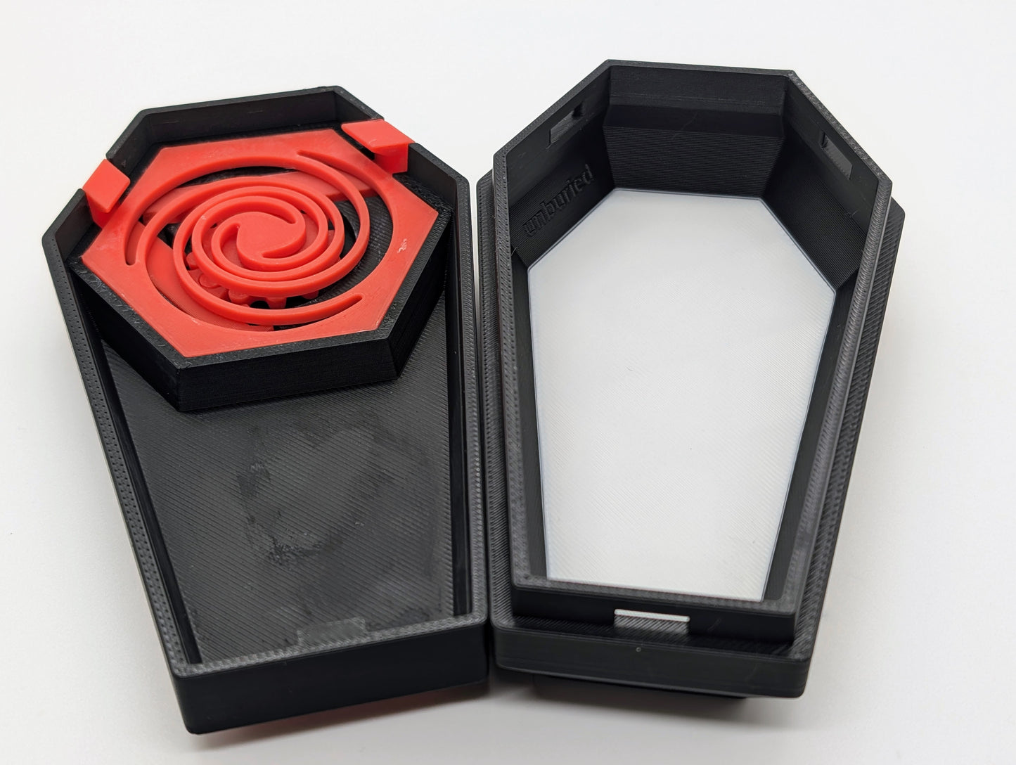 A photograph of a 3D-printed, coffin-shaped box with the lid and base shown side by side. The lid, on the left, features a red spiral design inset within a black border. The base, on the right, is open, revealing the word 'unburied' embossed on the interior wall near the top. The box has a modern, angular design with a smooth surface, and the contrast between the red and black elements adds visual interest. The white background highlights the details and craftsmanship of the 3D-printed object.