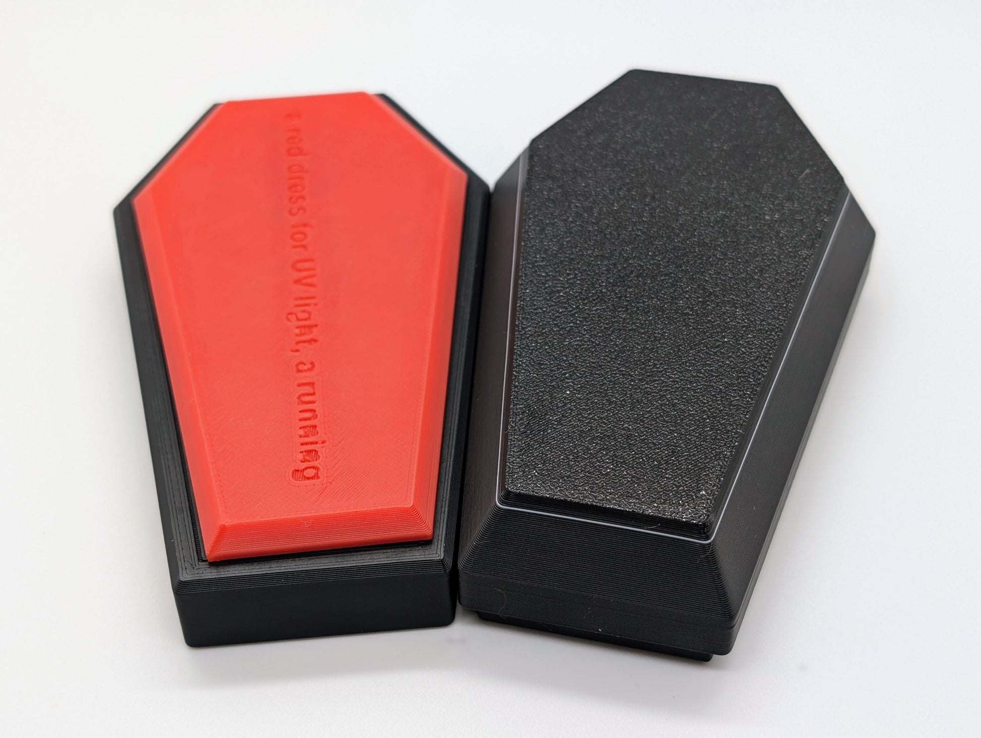 A photograph of two coffin-shaped 3D-printed objects placed side by side on a white background. The object on the left has a red face with embossed text that reads, 'a red dress for UV light, a running,' and a black border. The object on the right is entirely black with a textured surface, giving it a sleek and solid appearance. The contrast between the red and black elements highlights the design and craftsmanship of the 3D-printed items.