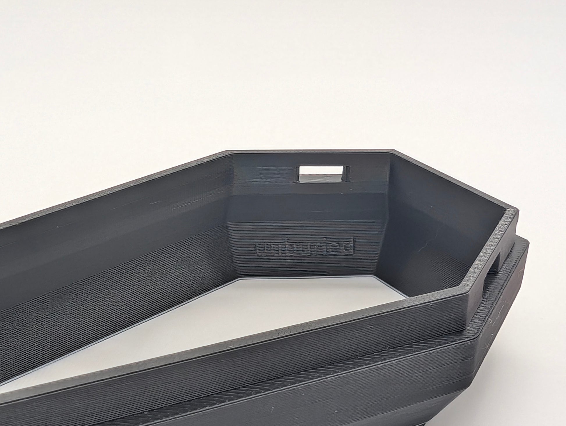 A close-up photograph of the interior of a black, 3D-printed coffin-shaped object. The inside of the object features the embossed word 'unburied' along one of the interior walls. The object is angular and has a modern, minimalist design, with clean lines and a smooth surface. The background is plain and white, allowing the details of the 3D print and the text to stand out clearly. The image emphasizes the craftsmanship and thoughtful design of the object.