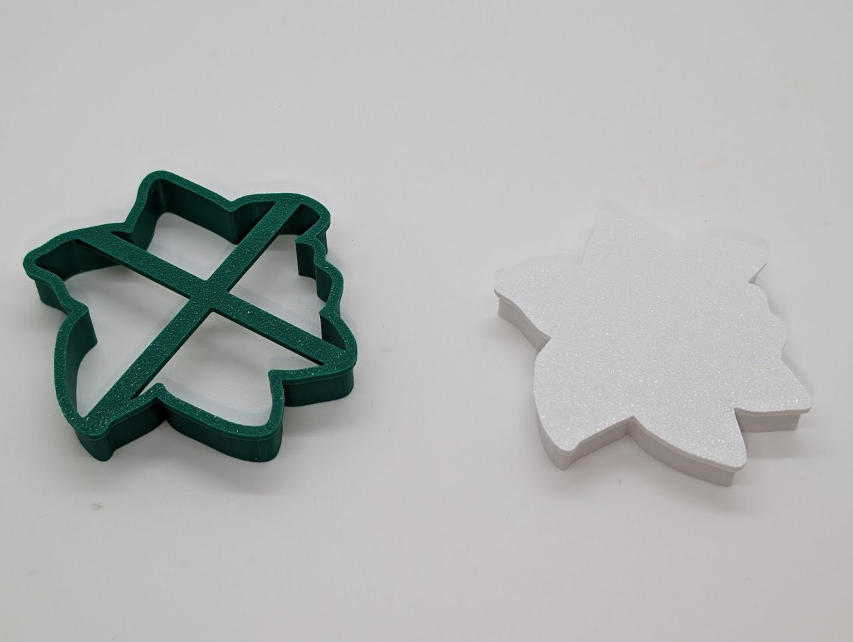 A photograph of two 3D-printed objects placed on a white surface. On the left is a green, flower-shaped cookie cutter with a cross pattern inside, designed to support the shape of the cutter. On the right is a solid white object, also shaped like a flower, which appears to be a complementary piece to the cutter. The simple background emphasizes the shapes and colors of the objects, highlighting their clean lines and the precision of the 3D printing.