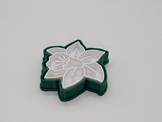 A close-up photograph of a 3D-printed cookie cutter shaped like a flower. The cookie cutter has a green outer edge with white inner details, which form the petals and center of the flower. The design is intricate, with varying depths to create a detailed floral pattern when used. The object is placed against a plain white background, highlighting the precision and craftsmanship of the 3D print.